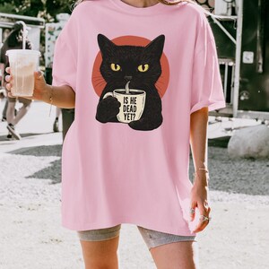May include: Pink t-shirt featuring a black cat illustration with yellow eyes, holding a mug. The mug has the text "IS HE DEAD YET?". The cat is set against a red circle. The shirt is a casual, oversized fit.