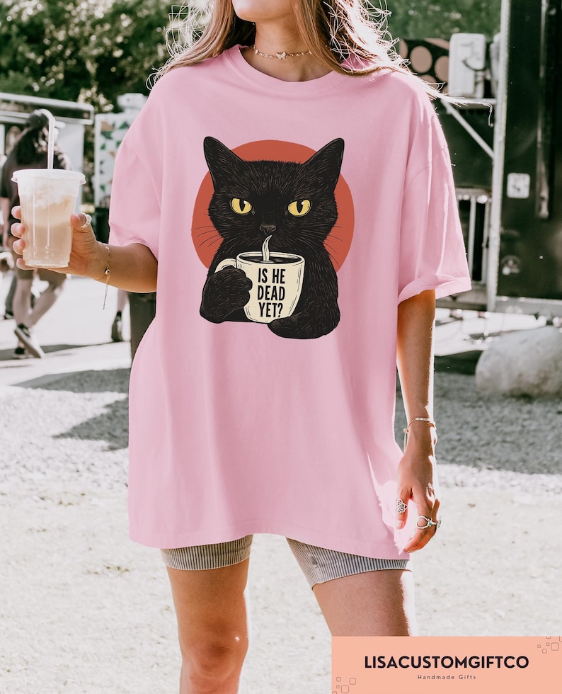 May include: Pink t-shirt featuring a black cat illustration with yellow eyes, holding a mug. The mug has the text "IS HE DEAD YET?". The cat is set against a red circle. The shirt is a casual, oversized fit.