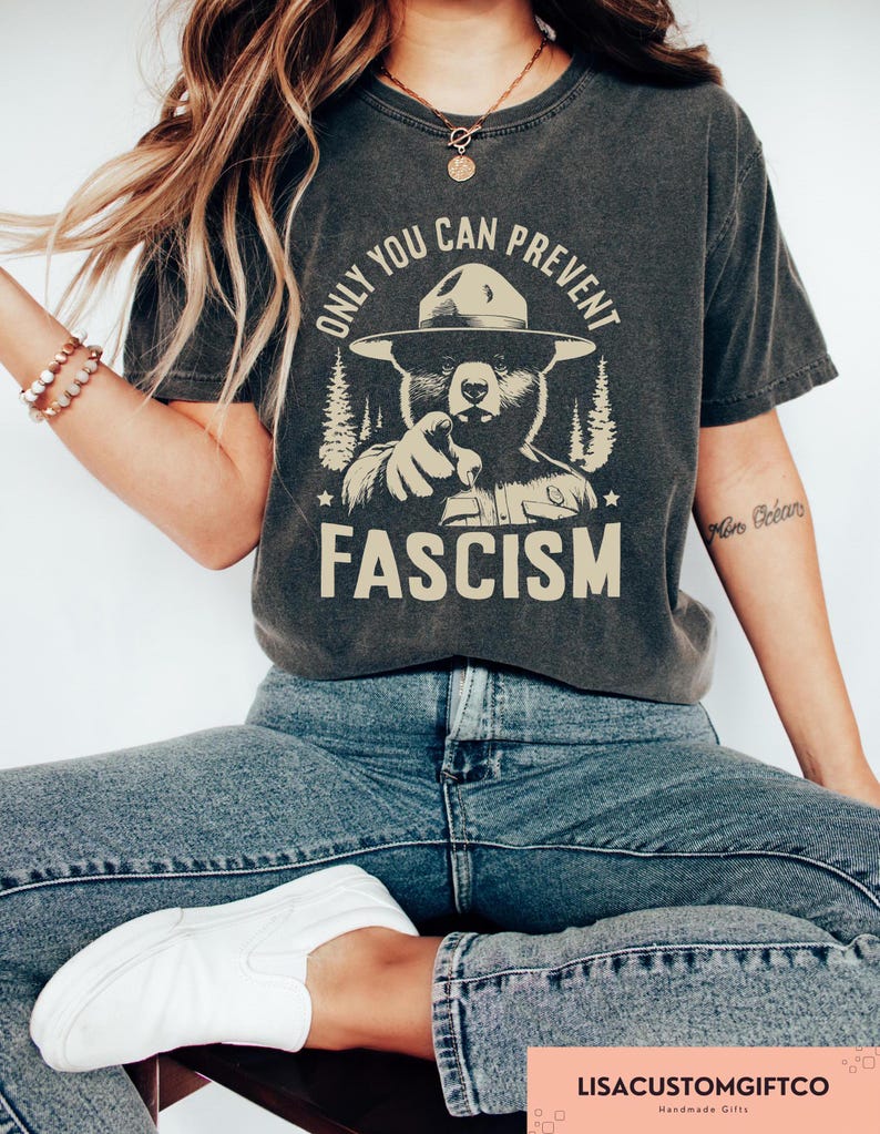 K&ouml;nnte beinhalten: Ein dunkelgraues T-Shirt mit einem Aufdruck eines B&auml;ren, der einen Rangerhut tr&auml;gt und auf den Betrachter zeigt. Der Text "Only you can prevent fascism" ist auf dem T-Shirt aufgedruckt.