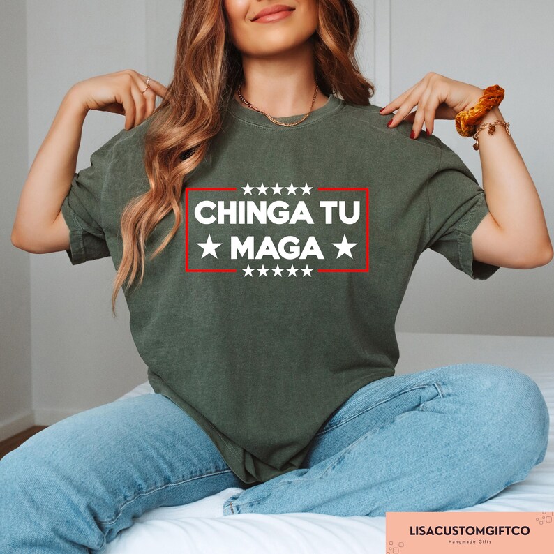 May include: An olive green t-shirt featuring the text "CHINGA TU MAGA" in white within a red rectangle, with white stars above and below. The shirt is worn with blue jeans.