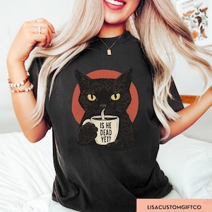 May include: Black t-shirt featuring a cartoon cat with yellow eyes holding a coffee mug. The mug has the text "IS HE DEAD YET?" printed on it. The cat is set against a red circle.