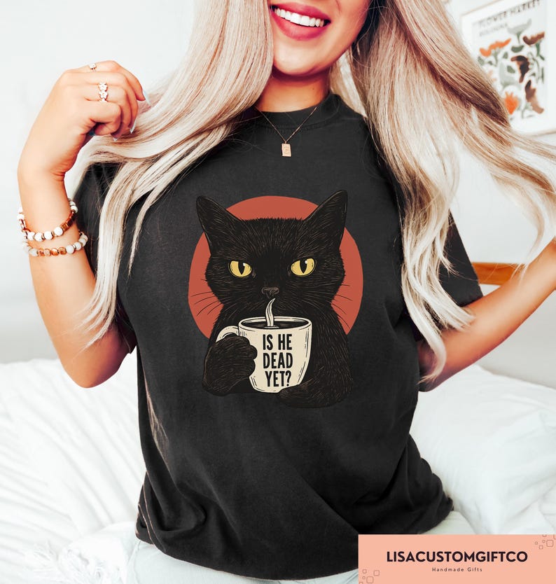 May include: Black t-shirt featuring a cartoon cat with yellow eyes holding a coffee mug. The mug has the text "IS HE DEAD YET?" printed on it. The cat is set against a red circle.