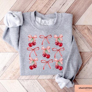 May include: A heather gray sweatshirt featuring a pattern of red cherries tied with pink bows. The cherries are arranged in a grid pattern across the front of the sweatshirt. The text "LISACUSTOMGIFTCO" is visible in the bottom right corner.