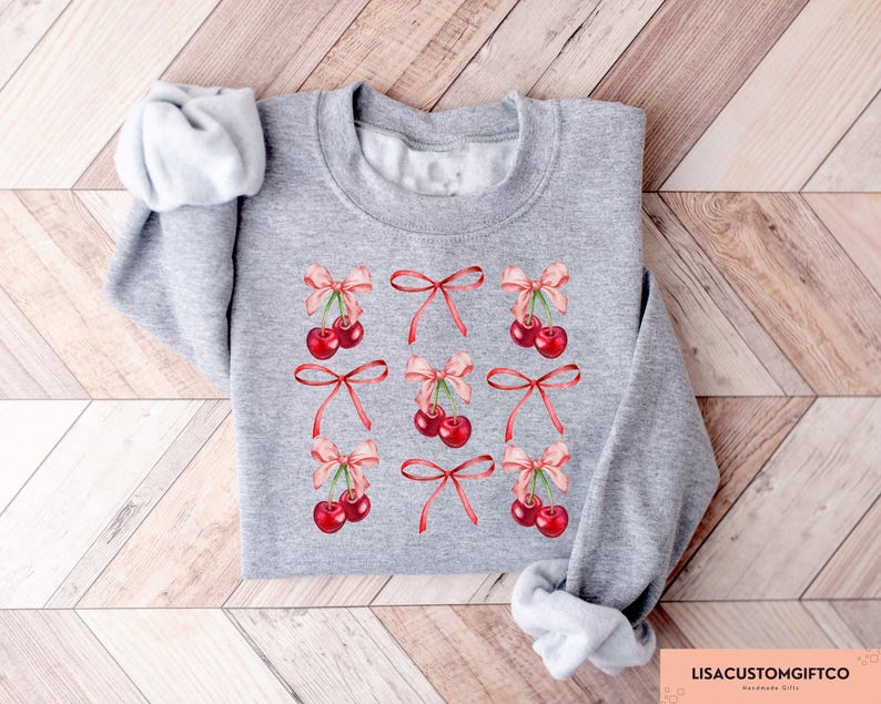 May include: A heather gray sweatshirt featuring a pattern of red cherries tied with pink bows. The cherries are arranged in a grid pattern across the front of the sweatshirt. The text "LISACUSTOMGIFTCO" is visible in the bottom right corner.