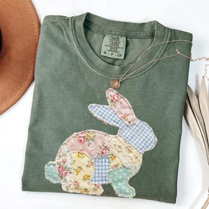 Può includere: T-shirt verde oliva con un design a coniglio patchwork. Il coniglio è realizzato con tessuti floreali, a quadri e a pois in colori pastello. Una collana d'argento con un piccolo ciondolo è appoggiata sulla maglietta. La maglietta è piegata su una superficie bianca.