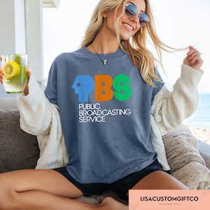 May include: A blue t-shirt featuring the PBS logo in blue, orange, and green. The words "PUBLIC BROADCASTING SERVICE" are printed below the logo in white. The shirt is being worn.