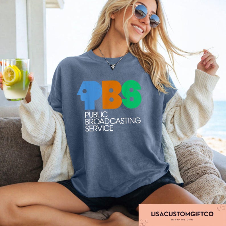 May include: A blue t-shirt featuring the PBS logo in blue, orange, and green. The words "PUBLIC BROADCASTING SERVICE" are printed below the logo in white. The shirt is being worn.