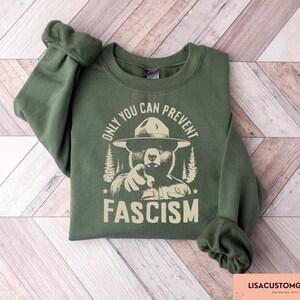 K&ouml;nnte beinhalten: Gr&uuml;nes Sweatshirt mit einem Aufdruck eines B&auml;ren, der einen Rangerhut tr&auml;gt. Der Text "Only You Can Prevent Fascism" ist auf dem Sweatshirt aufgedruckt.