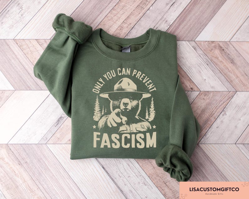 K&ouml;nnte beinhalten: Gr&uuml;nes Sweatshirt mit einem Aufdruck eines B&auml;ren, der einen Rangerhut tr&auml;gt. Der Text "Only You Can Prevent Fascism" ist auf dem Sweatshirt aufgedruckt.