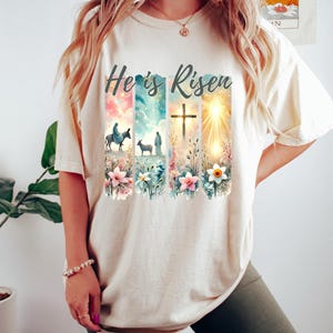 May include: A beige t-shirt with the text "He is Risen" printed on the front. The design features a watercolor style image of a cross, a sunrise, and two figures walking with a donkey and a sheep. The design is surrounded by flowers.