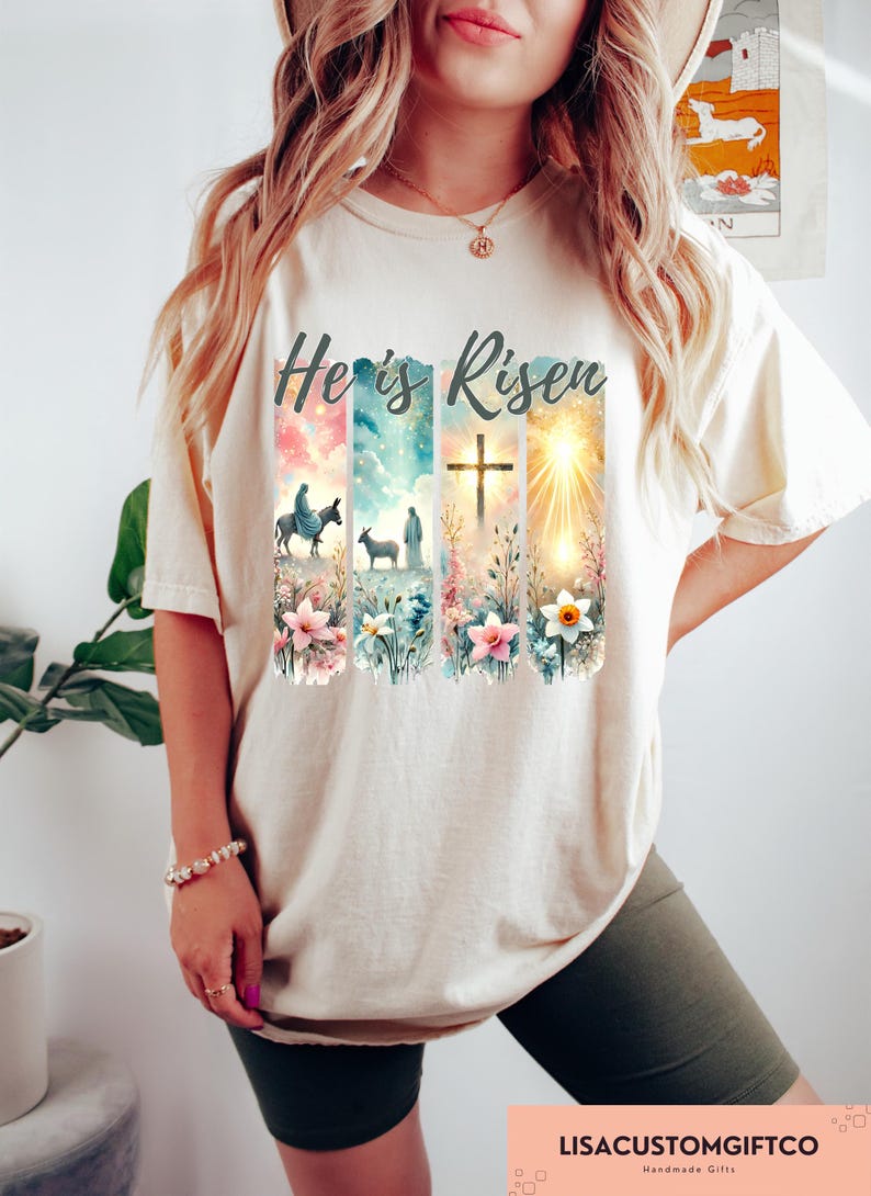 May include: A beige t-shirt with the text "He is Risen" printed on the front. The design features a watercolor style image of a cross, a sunrise, and two figures walking with a donkey and a sheep. The design is surrounded by flowers.
