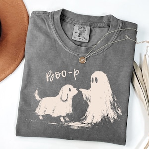 以下が含まれることがあります： チャコールグレーの半袖Tシャツ。犬と幽霊が向かい合っているライトクリーム色のグラフィックと、その上に「BOO-p」という文字がプリントされています。クルーネックで、襟の内側にはComfort Colorsのブランドラベルが付いています。