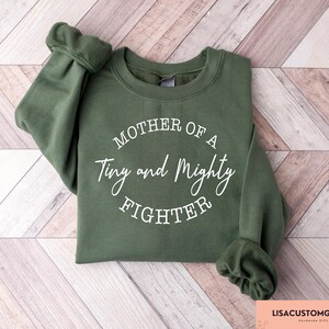 May include: A green sweatshirt with the text "Mother of a Tiny and Mighty Fighter" printed in white on the front.