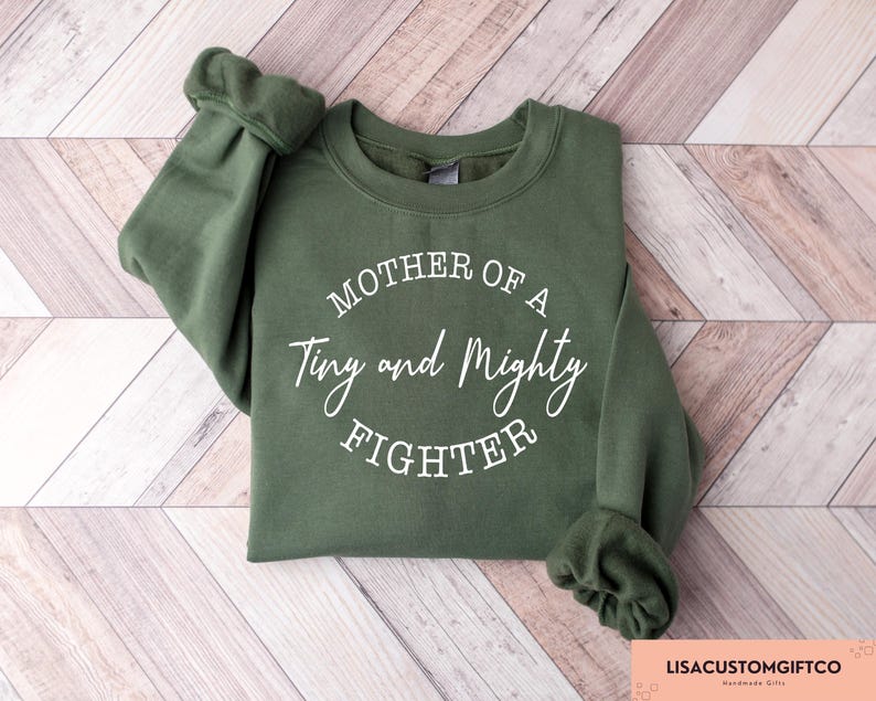 May include: A green sweatshirt with the text "Mother of a Tiny and Mighty Fighter" printed in white on the front.