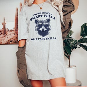 May include: A light gray t-shirt with the text "YOU'RE EITHER A SMART FELLA OR A FART SMELLA" and a raccoon wearing sunglasses graphic in navy blue. The shirt is a casual, oversized fit.