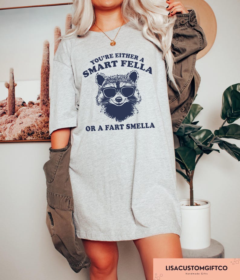 May include: A light gray t-shirt with the text "YOU'RE EITHER A SMART FELLA OR A FART SMELLA" and a raccoon wearing sunglasses graphic in navy blue. The shirt is a casual, oversized fit.