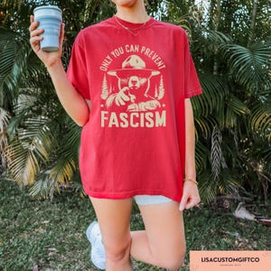 K&ouml;nnte beinhalten: Rotes T-Shirt mit einem Cartoon-B&auml;ren, der einen Rangerhut tr&auml;gt und auf den Betrachter zeigt. Der Text "Only you can prevent fascism" ist auf dem T-Shirt aufgedruckt.