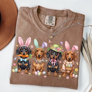 May include: Brown Comfort Colors t-shirt featuring a graphic of four dachshunds dressed as Easter bunnies. Each dog wears bunny ears and a unique accessory, such as a carrot, hat, or scarf. The shirt has a crew neck and short sleeves.