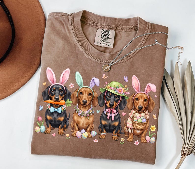 May include: Brown Comfort Colors t-shirt featuring a graphic of four dachshunds dressed as Easter bunnies. Each dog wears bunny ears and a unique accessory, such as a carrot, hat, or scarf. The shirt has a crew neck and short sleeves.