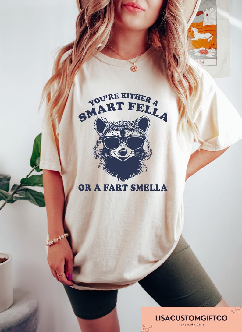 May include: Cream-colored t-shirt with a raccoon wearing sunglasses graphic. The text reads "You're either a Smart Fella or a Fart Smella." The shirt is part of a handmade gifts collection.