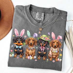 May include: A gray Comfort Colors t-shirt featuring four dachshunds dressed as Easter bunnies. Each dog wears bunny ears, a floral collar, and holds Easter eggs or a carrot. A necklace and a brown hat are also visible.