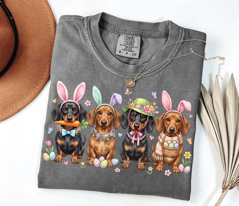 May include: A gray Comfort Colors t-shirt featuring four dachshunds dressed as Easter bunnies. Each dog wears bunny ears, a floral collar, and holds Easter eggs or a carrot. A necklace and a brown hat are also visible.