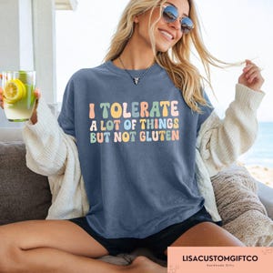 K&ouml;nnte beinhalten: Eine Frau tr&auml;gt ein blaues T-Shirt mit dem Text "I tolerate a lot of things but not gluten" in bunten Buchstaben. Sie sitzt auf einem Sofa und h&auml;lt ein Glas Limonade.