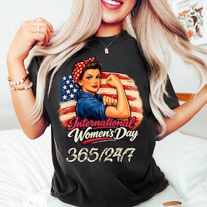 May include: Black t-shirt featuring a graphic of a woman flexing her arm in front of an American flag, with the text "International Women's Day" and "365/24/7". The woman is wearing a red bandana and a blue shirt.