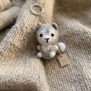 May include: A small, crocheted gray and white husky dog keychain with black bead eyes and a stitched nose. The plush figure features a metal keyring attached to the top and a small, square tan paper tag with the letter W printed on it.