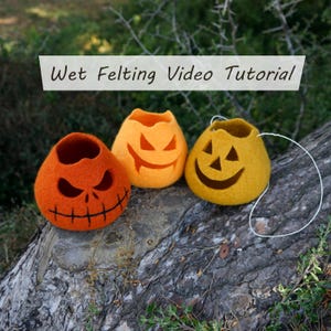 May include: Three felted pumpkin-shaped decorations in orange and yellow, each with a carved jack-o'-lantern face. The pumpkins are displayed on a weathered log, with the text "Wet Felting Video Tutorial" in the background.