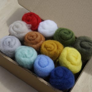 May include: A selection of twelve colourful wool felt balls in a cardboard box. The balls are in shades of red, white, grey, brown, green, yellow, blue, and teal. The felt balls are neatly arranged, perfect for various craft projects.