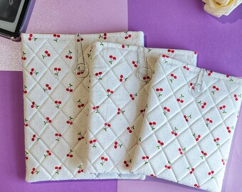 Puede incluir: Tres fundas de tela acolchada blanca con un estampado de cerezas, sujetas con clips plateados. Las fundas están dispuestas sobre una superficie morada y lavanda. Un dispositivo Kindle es visible en la esquina superior izquierda.