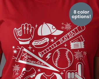 May include: A red t-shirt featuring a white graphic print of baseball-themed icons including a glove, cap, hot dog, bat with the text PLAY BALL!, tickets, a Los Angeles pennant, a ball, a cleat, a jersey with LET'S GO HALOS, and a foam finger.