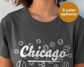 May include: A dark gray short-sleeve t-shirt featuring the word Chicago in white script lettering, underlined by a horizontal line, surrounded by multiple white baseball graphics. A circular orange badge states 5 color options.