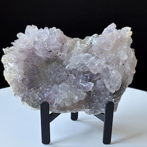 May include: A cluster of pale purple amethyst crystals forming a jagged, irregular shape. The mineral specimen features numerous translucent, pointed crystal points and is displayed on a black metal stand with three vertical prongs.