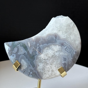 Puede incluir: Una rebanada de ágata en forma de media luna con un patrón blanco, gris y verde, exhibida en un soporte de metal dorado.