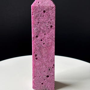 May include: A pink crystal obelisk with a rectangular shape and a pointed top. The crystal has a porous texture with small, dark holes. It stands on a white surface against a black backdrop. A decorative item.
