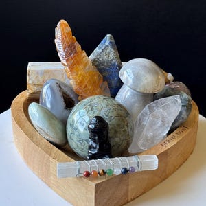 May include: A heart-shaped wooden bowl filled with various crystals, including a carved feather, a mushroom, and a small Buddha statue. A selenite wand with colorful beads is in the foreground. The words "prayer bowl" are etched on the side.