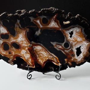 May include: A polished agate slice featuring deep black, translucent amber, and white crystalline patterns. The stone has a rough, dark outer crust and is displayed on a black metal wire stand with curled feet, showcasing natural mineral formations.