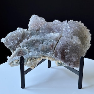 May include: A large, jagged amethyst crystal cluster with deep purple and translucent crystalline points, resting on a black metal display stand. The mineral specimen features a rough, light-colored base and a dense formation of sparkling points.