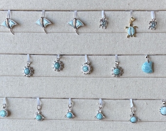 Puede incluir: Una colección de colgantes de plata con incrustaciones de piedras azul claro. Los diseños incluyen mantarrayas, tortugas y formas redondas y de sol. Los colgantes se exhiben sobre un fondo neutro.