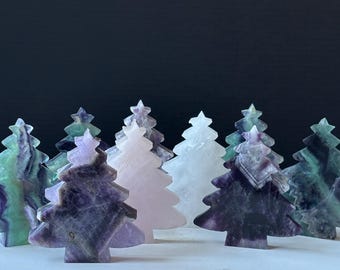 May include: A collection of carved gemstone Christmas trees in various colors, including green, purple, and white. The trees are arranged in a row, each with a star on top. The trees are made of fluorite and quartz.