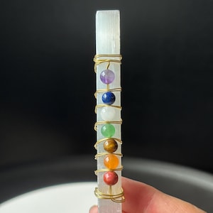 Pode incluir: Uma varinha de selenita retangular transparente adornada com sete contas de pedras preciosas coloridas, envoltas com arame dourado. As contas estão dispostas verticalmente, mostrando um espectro de cores, incluindo vermelho, laranja, amarelo, verde, branco, azul e roxo.