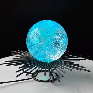 May include: A blue illuminated orb lamp with a textured surface, set on a black metal starburst base. The lamp emits a bright, cool light, creating a modern aesthetic. The base has multiple black rods extending outwards.