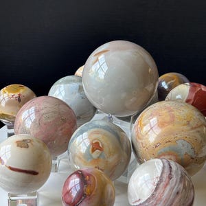 May include: A selection of polished stone spheres in shades of cream, pink, brown, and grey. Each sphere is presented on a clear acrylic stand, highlighting the natural patterns and textures. The spheres are of varying sizes, creating a diverse display.