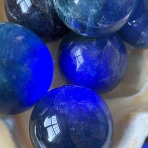 Puede incluir: Un conjunto de esferas pulidas de color azul oscuro, posiblemente piedras preciosas o bolas de cristal decorativas. Las esferas var&iacute;an ligeramente en tonalidad, algunas parecen m&aacute;s oscuras y otras m&aacute;s claras, reflejando la luz. Descansan en un cuenco blanco texturizado.