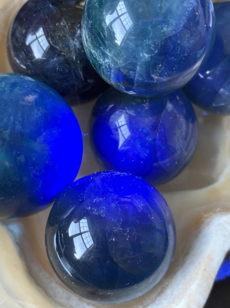 Puede incluir: Un conjunto de esferas pulidas de color azul oscuro, posiblemente piedras preciosas o bolas de cristal decorativas. Las esferas var&iacute;an ligeramente en tonalidad, algunas parecen m&aacute;s oscuras y otras m&aacute;s claras, reflejando la luz. Descansan en un cuenco blanco texturizado.