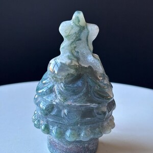 May include: A carved gemstone figurine in the shape of a Christmas tree. The tree is primarily green and white with a star on top. The base of the tree is a darker shade of green and purple.