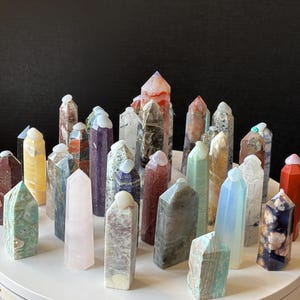 May include: A selection of multi-coloured crystal points, each with a unique shape and colour. The crystals are arranged on a white surface against a dark background. Colours include pink, green, yellow, and blue.