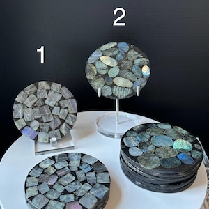 May include: Set of four round coasters made of iridescent labradorite stone. The coasters are black with blue, green, and purple iridescent accents. The coasters are arranged in a circle on a white surface.